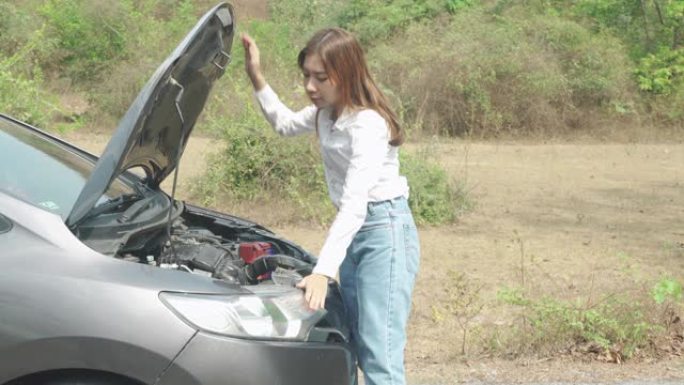 亚洲年轻不快乐的女人在乡村道路上的敞篷抛锚汽车前检查损坏的汽车发动机，等待道路援助服务。旅途中的破车