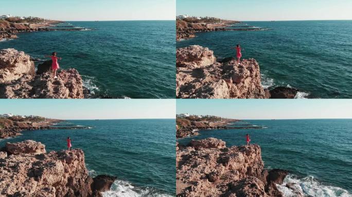 穿着红色连衣裙的女孩在强劲的海风中拍打，站在山崖的边缘，双臂仰望地平线。在塞浦路斯的岩石悬崖上，双臂