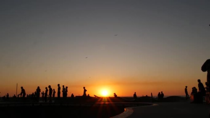 年轻跳跃滑板手骑长板的剪影，夏天的日落背景。加州洛杉矶威尼斯海洋海滩滑板公园。极限公园滑板坡道上的青