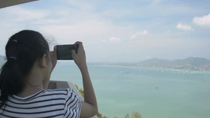 美丽的女孩拿着手机在观景台上拍照时的后视，海湾风景优美，天空多云。