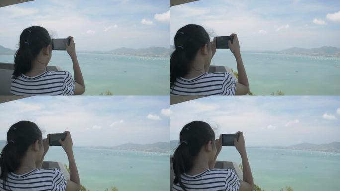 美丽的女孩拿着手机在观景台上拍照时的后视，海湾风景优美，天空多云。
