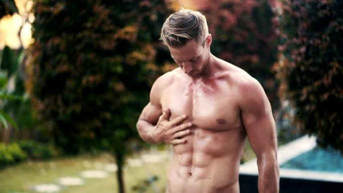 运动性感的男子应用日光浴油在他的身体与泳池的背景