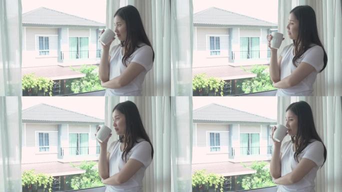 美丽的亚洲女人醒来在她的床上充分休息，打开窗台上的窗帘，看着窗外的一杯咖啡，以获取新鲜的空气，在阳光