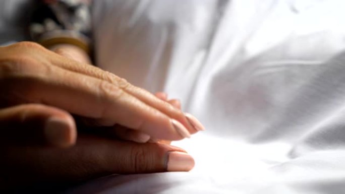 无法辨认的女人握住并轻轻地抚摸母亲的手，表现出关怀或爱心。女儿安慰躺在诊所床上的年迈妈妈的皱纹手臂。