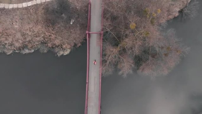 跑过桥的女人的空中俯视图。穿着鲜艳夹克的运动型女运动员在城市公园的过河桥上奔跑。有动力的年轻女孩在户