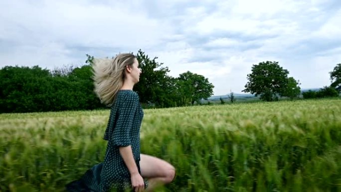 年轻快乐的金发女孩在傍晚的雨中天空背景下在绿色的麦田上奔跑。侧视图