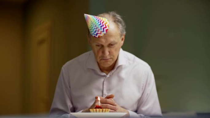 缩小坐在家里餐桌旁，低头看着蜡烛燃烧的纸杯蛋糕的沮丧老人，在生日那天感到不开心