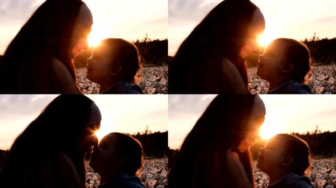 日落时，一个年轻女子亲吻她的小儿子坐在花草地上的特写剪影。