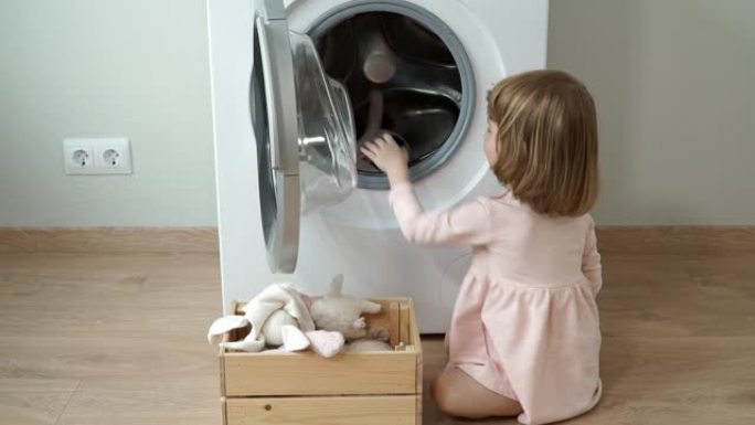 微笑可爱的3岁小女孩的肖像坐在层压地板上，打开舱门，放入洗衣机玩具进行洗涤