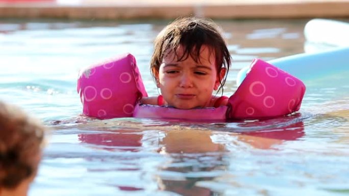 心烦意乱的小女孩在游泳池水中蹒跚学步，抱怨穿着漂浮物