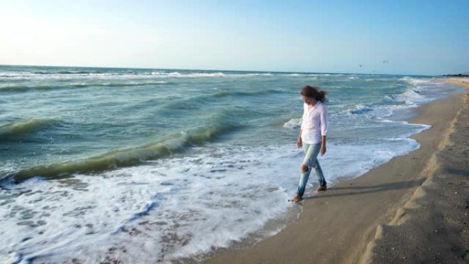 穿着牛仔裤和白色上衣的女孩在大风大浪的夏日沿着海边散步