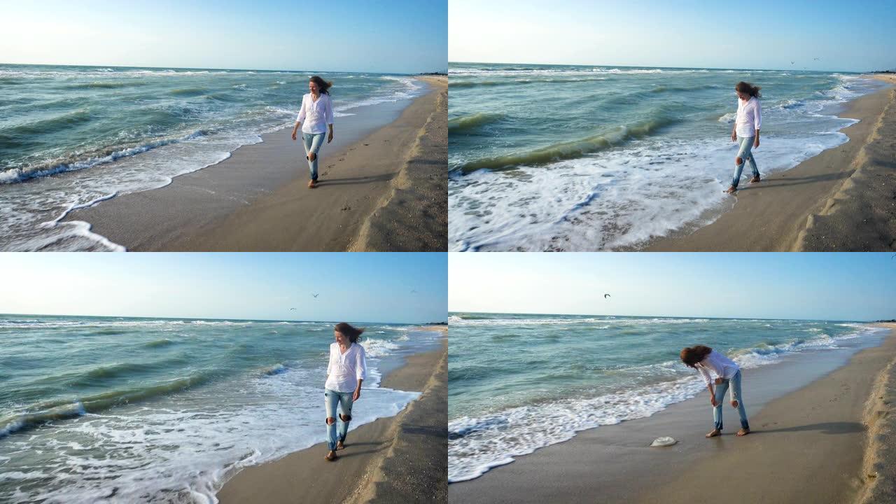 穿着牛仔裤和白色上衣的女孩在大风大浪的夏日沿着海边散步