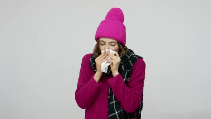 冬天不健康的女孩帽子和围巾在餐巾纸上打喷嚏，看起来带着不高兴的恶心表情