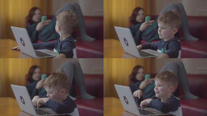 金发碧眼的学龄前男孩在笔记本电脑上打字，微笑着看着电脑屏幕。背景是手机躺在沙发上的母亲。孩子坐在桌旁