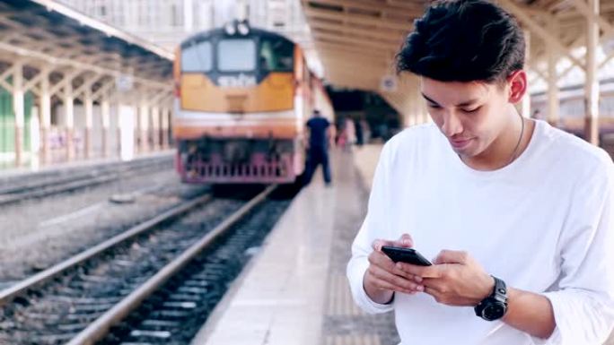 中等拍摄的潮人在火车站等火车，用他的手机，摇头，与火车在背景，在曼谷。亚洲人。