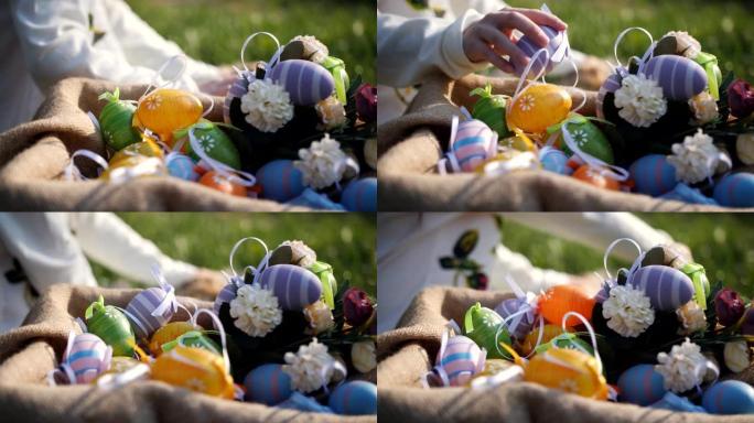 女孩将散落在草地上的彩色复活节彩蛋添加到篮子中，并抚摸着蓬松的灰色兔子，后者坐在她旁边的格子毯子上。