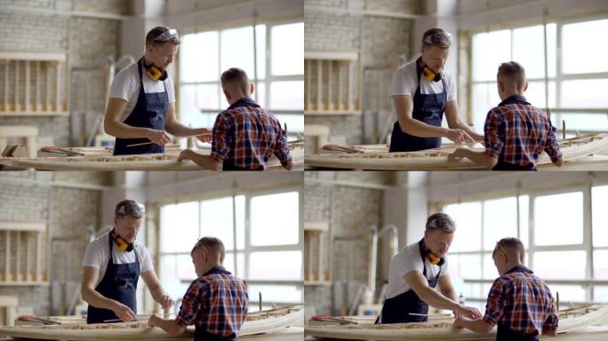 围裙中的中年父亲和小儿子一起在木工车间的工作台上建造小木船