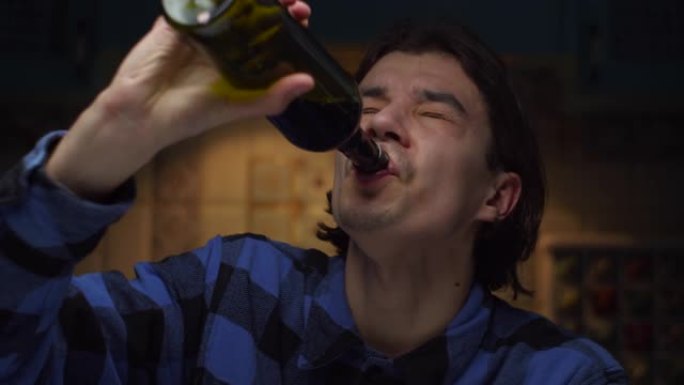 沮丧的20多岁年轻人喝瓶装酒的特写肖像。穿着蓝色衬衫的男人喝酒，拳头窒息。