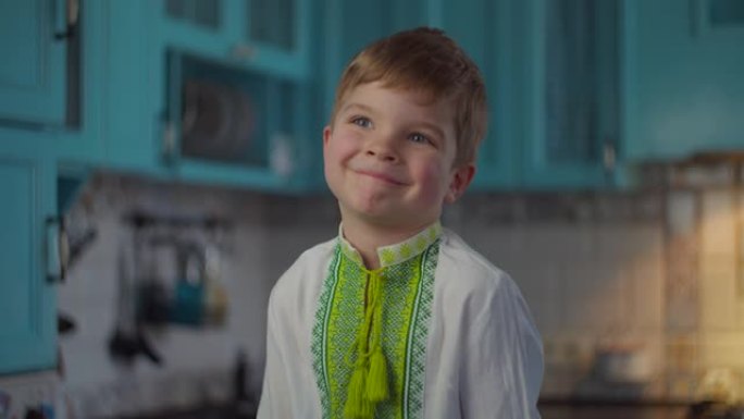 穿着刺绣民族服装的金发男孩在蓝色厨房里微笑着展示着自己的情感。穿着白色针织衬衫的孩子，慢动作中有民族