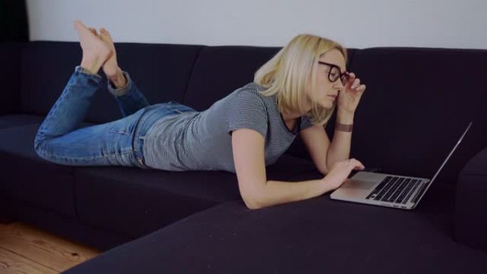 随意的女人，金发和眼镜躺在黑色沙发上，使用笔记本电脑客厅。