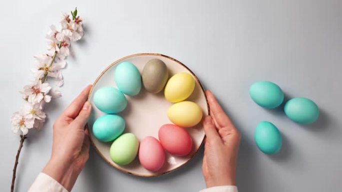 双手将装有彩色鸡蛋的盘子放在复活节装饰的桌子上。带有花朵的树枝和蓝色背景
