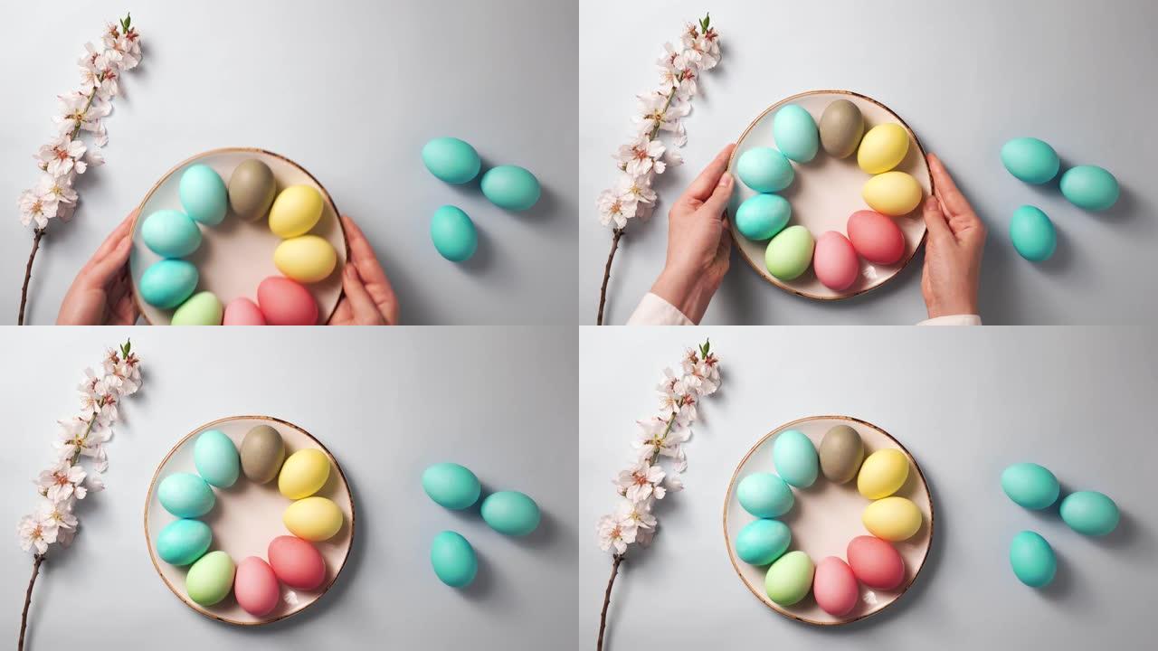 双手将装有彩色鸡蛋的盘子放在复活节装饰的桌子上。带有花朵的树枝和蓝色背景