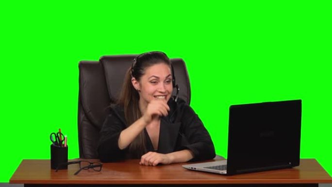 戴着耳机的女商人用网络摄像头说话，带着喜悦的心情看着笔记本电脑。绿屏。慢动作
