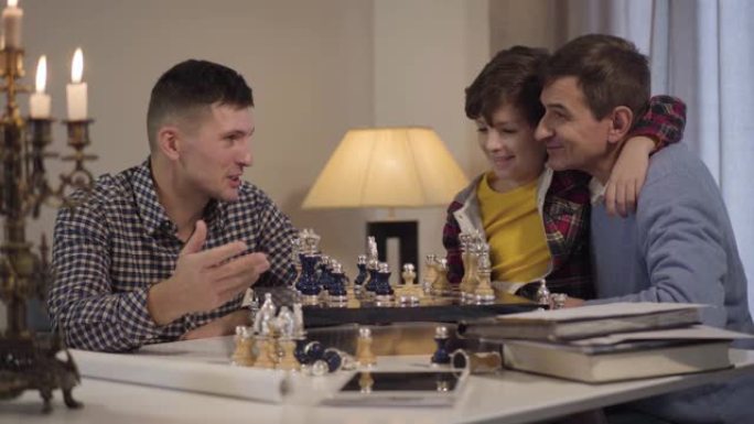 快乐的高加索祖父和孙子对男孩的父亲下棋。微笑的家人在家里共度夜晚。休闲，生活方式，幸福