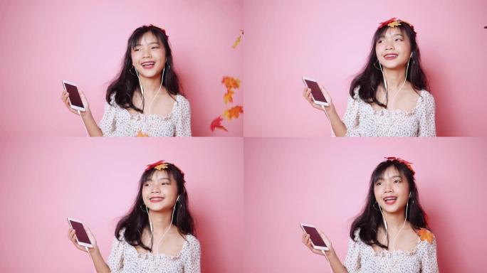 快乐的亚洲少女戴着耳机，在粉色背景上玩掉落的枫叶。慢动作。