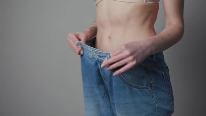 女人显示她的体重减轻，穿着她的旧牛仔裤。穿着大牛仔裤的苗条女孩展示了她开始吃健康食物时是如何减肥的