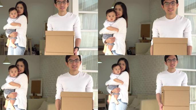 快乐的亚洲父母抱着可爱的婴儿和箱子，带着物品看着相机，同时搬进新房子。年轻夫妇家庭搬迁