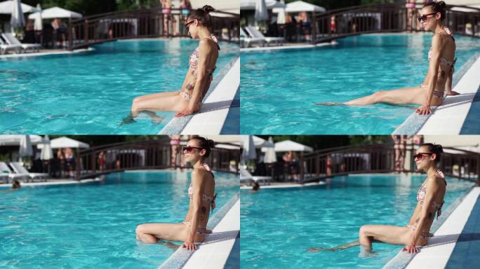 年轻匀称的女人，腹部平坦，坐在泳池边缘，双腿浸入水中，享受阳光明媚的日子