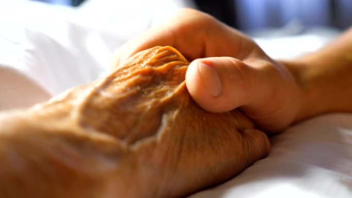 忧心忡忡的年轻人握着并轻轻地抚摸着生病的祖母的手，表现出关心或爱。儿子安慰躺在床上的年迈妈妈皱巴巴的