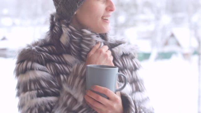 穿着皮大衣和针织毛线帽的快乐美丽女孩在一个寒冷的冬天早晨从杯子里喝咖啡。一名年轻女子在街上喝茶，下雪
