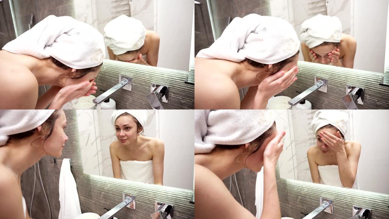 一名妇女站在水槽附近的浴室里，头上穿着白毛巾，用水龙头里的水洗脸。看着镜子的倒影和微笑