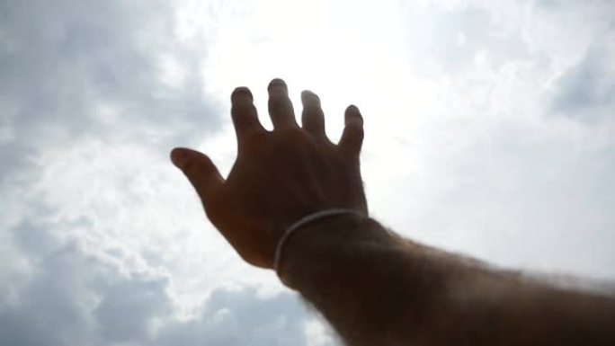 在多云的天空背景下，明亮的阳光穿透年轻人伸出手臂的手指。雄性手的特写伸向太阳。暑假或假期的概念。慢动