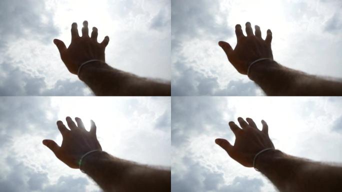 在多云的天空背景下，明亮的阳光穿透年轻人伸出手臂的手指。雄性手的特写伸向太阳。暑假或假期的概念。慢动