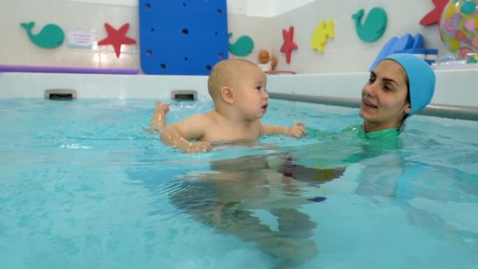一个快乐的幼儿在儿童游泳池与教练一起学习游泳和游泳的特写镜头。他环顾四周，微笑着。婴儿游泳。概念