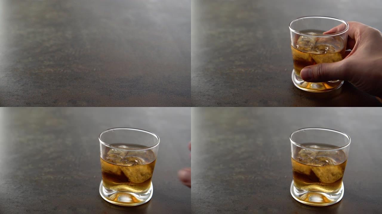 男人喝一杯加冰块的陈年金威士忌，然后放在桌子上。用手将琥珀色酒精饮料和岩石放在岩石背景上