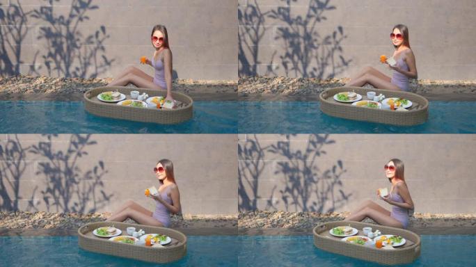 美丽的年轻亚洲女人快乐的微笑享受早餐漂浮在室外游泳池周围
