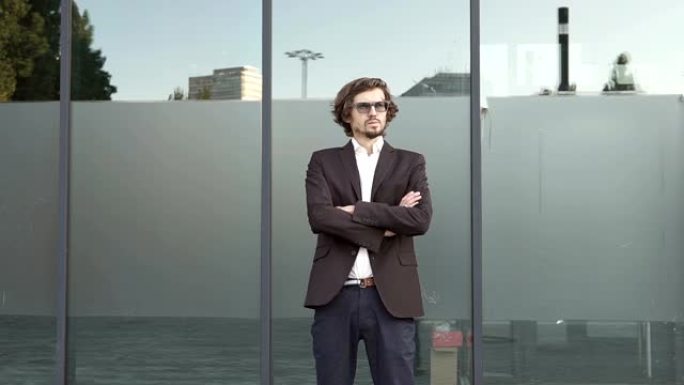 穿着西装的男人双臂交叉在胸前，现代玻璃办公楼背景。一个男人看着镜头，骄傲而自信。