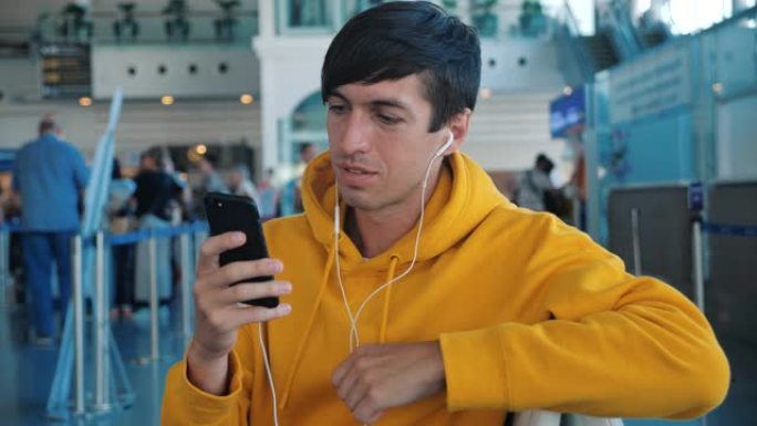 男子坐在机场休息室的长椅上，一边看着智能手机的屏幕，一边通过耳机听音乐。穿着黄色连帽衫的男性潮人在机