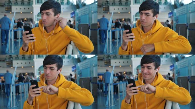 男子坐在机场休息室的长椅上，一边看着智能手机的屏幕，一边通过耳机听音乐。穿着黄色连帽衫的男性潮人在机