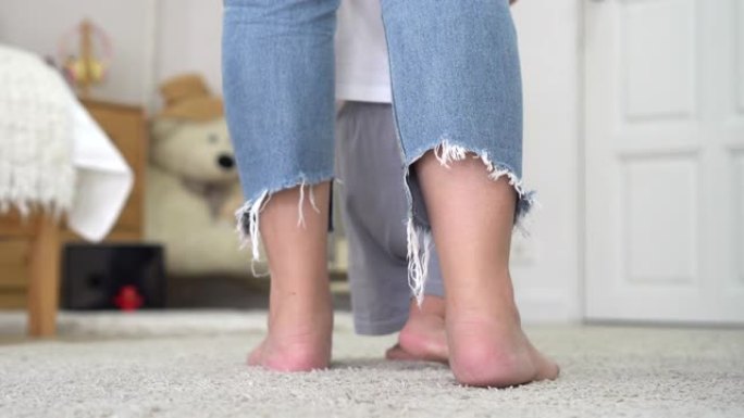穿着牛仔裤的赤脚女人在家里舒适的托儿所教小婴儿在柔软的白色地毯上行走