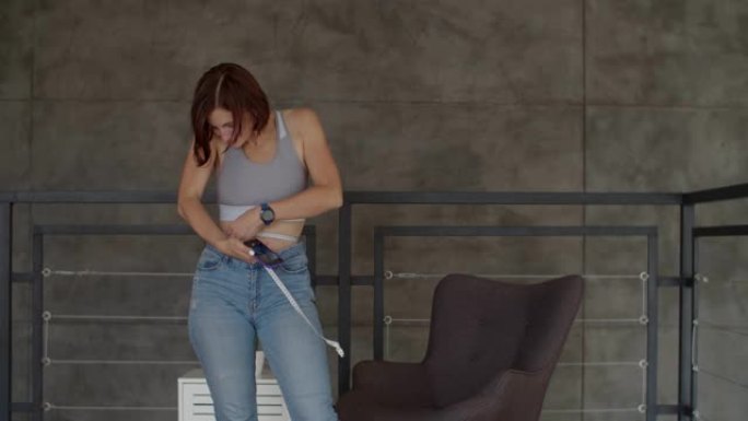 年轻女子在卧室穿着旧牛仔裤时欣赏减肥的结果。微笑的女性用厘米胶带测量她的腰部。
