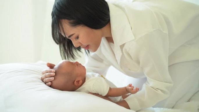 穿着白色衣服的年轻亚洲妈妈抱着自己的婴儿在家里亲吻新生儿子的侧视图