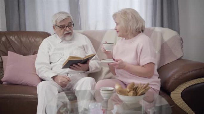 快乐的白人高级夫妇在家度过平静的夜晚。英俊的成熟男人看书，他美丽的老伴喝茶。已婚退休人员在室内休息。