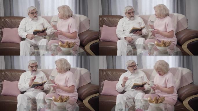 快乐的白人高级夫妇在家度过平静的夜晚。英俊的成熟男人看书，他美丽的老伴喝茶。已婚退休人员在室内休息。