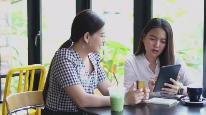 两名亚洲妇女在咖啡馆喝咖啡，并在智能手机上在线购物