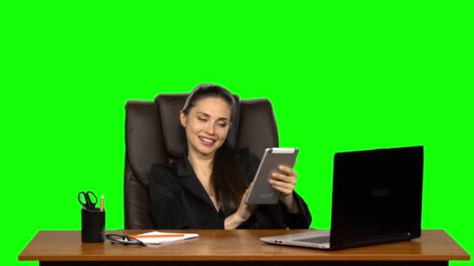 工人女孩坐在工作场所，靠在皮椅上，在平板电脑上交流，撒娇地微笑。绿屏